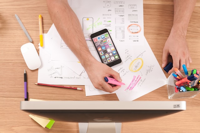 Overhead shot of someone's arms working at a desk with paper, a phone, and a desktop computer